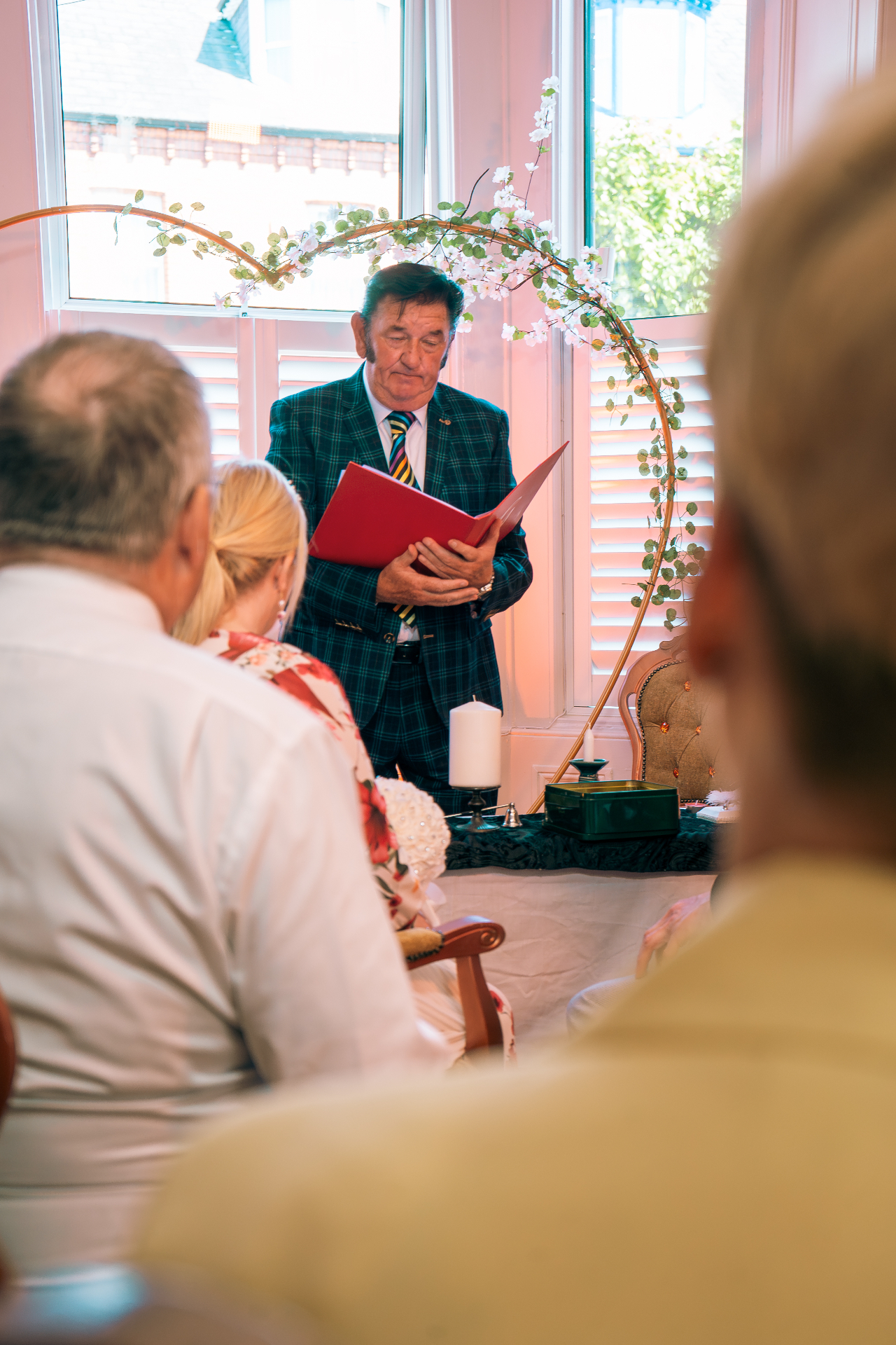 Greg Traynor officiating a wedding ceremony, reading from the red ceremony folder beneath a floral arch