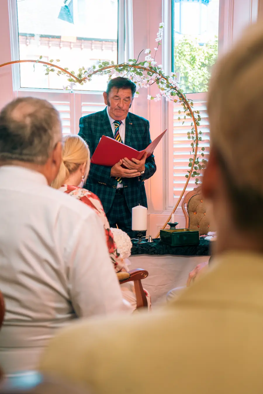 Greg Traynor officiating a wedding ceremony, reading from the red ceremony folder beneath a floral arch